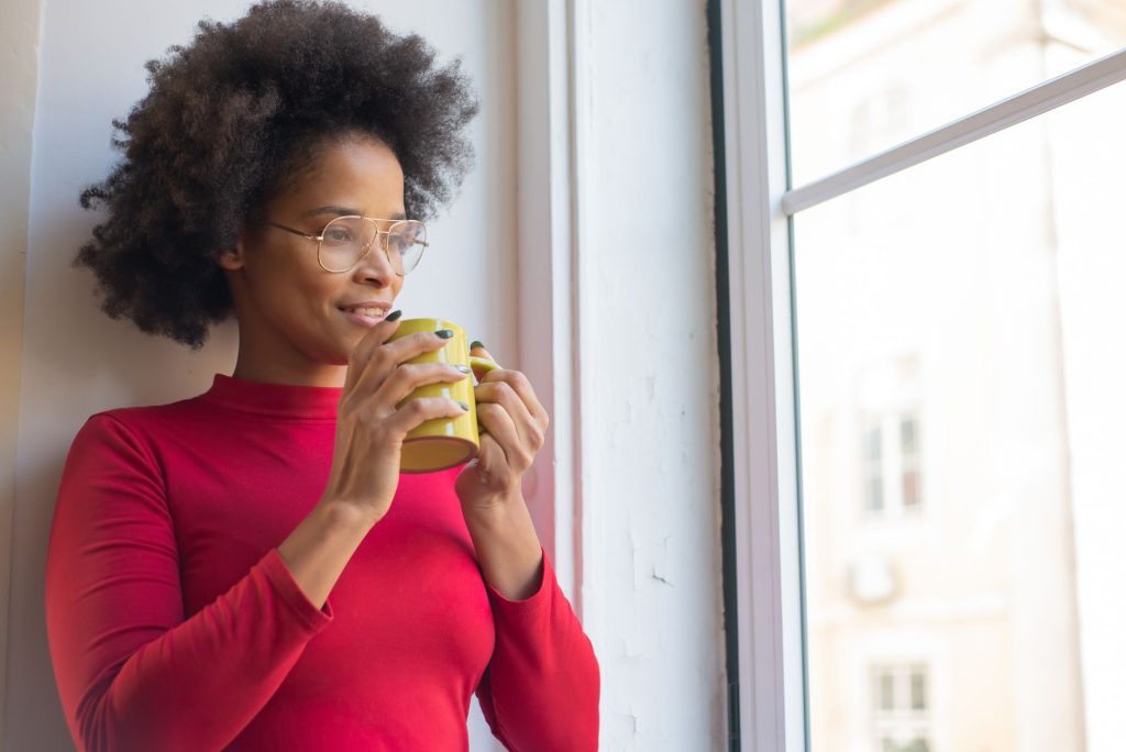Femme assise près d’une fenêtre avec un café, réfléchissant avant d’écrire